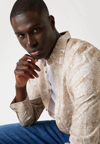 Beige gestreepte shirt gemaakt van lichtgewicht stof, met een button-down kraag, ingewikkelde ontwerpen en opgerolde mouwen. Draag het over een wit T-shirt.
