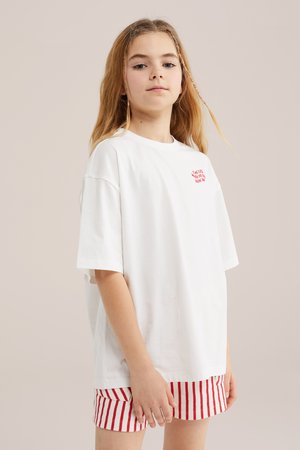 Fiatal, hosszú szőke hajú személy, aki túlméretezett fehér pólót és piros-fehér csíkos rövidnadrágot visel, egyszínű háttér előtt áll.