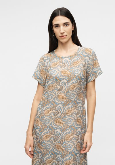 Γυναίκα με ίσια σκούρα μαλλιά που φοράει φόρεμα με κοντά μανίκια και περίπλοκο μπλε και καφέ μοτίβο πεϊσλί, στέκεται μπροστά σε απλό ανοιχτό φόντο.