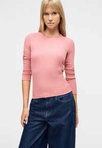 Νεαρή γυναίκα με μακριά ξανθά μαλλιά που φορεί ριμπ ροζ μπλούζα μακρυμάνικη και άνετο σκούρο μπλε τζιν, σε απλό φόντο.