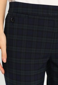 Marinblå rutiga byxor med texturerad tygkvalitet, slät front och dekorativ knappdetalj vid midjan.