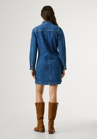 Robe en denim à manches longues, avec un col et une silhouette ajustée, présentant un ourlet brut. Associée à des bottes en suède marron jusqu'aux genoux.
