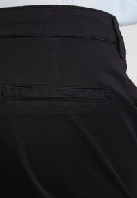 Gros plan sur l'arrière d'un pantalon noir présentant une poche horizontale avec des coutures soignées et une passante de ceinture à la taille.