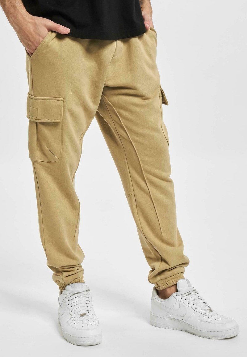 Homme portant un pantalon jogger cargo beige avec des poches latérales et des baskets blanches, debout les mains dans les poches sur fond blanc.