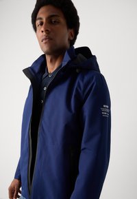 Veste étanche navy avec fermeture éclair à l'avant, capuche réglable, deux poches latérales et logo sur la manche. Texture lisse, design léger.