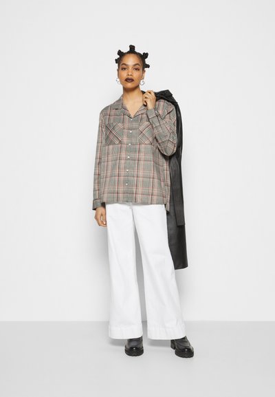 Camisa de botão xadrez em cinza com detalhes em vermelho, com duas bolsos no peito, combinada com calças largas brancas e botas pretas robustas, segurando um casaco preto.