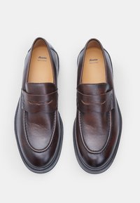 Mocassins en cuir marron avec un bout arrondi, des détails cousus et une sangle décorative sur le dessus. Présente une finition lisse et une semelle en caoutchouc.
