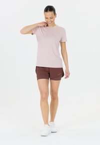 Rosa kurzärmliges T-Shirt, kombiniert mit braunen Sportshorts. Das Model trägt weiße Sneakers mit einem schlichten Design und glatten Texturen.