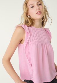 Rosa ærmeløs top med voluminøse skuldre, strukturerede lodrette plisseringer foran og en løs pasform. Stoffet virker let og blødt.