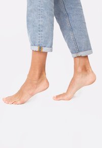 Braccialetto d'oro alla caviglia su un piede nudo, con jeans in denim azzurro chiaro arrotolati che mostrano un orlo sfilacciato e dettagli di cucitura a contrasto.