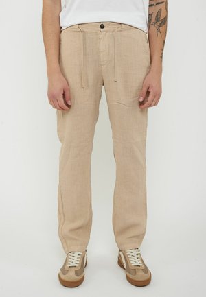 Uomo che indossa pantaloni di lino beige con coulisse e bottone, sneaker bianche e maglietta bianca, in piedi con le mani rilassate lungo i fianchi.