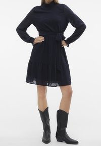 Marinblå klänning med texturerad tyg, långa ärmar, ett knytbälte vid midjan och en volang nederkant. Kombinationen med svarta cowboystövlar i ankellängd.