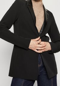 Černý blazer s ostrým klopem, přiléhavý střih a dlouhé rukávy. V kombinaci se světle hnědým rolákem a tmavými džínami.