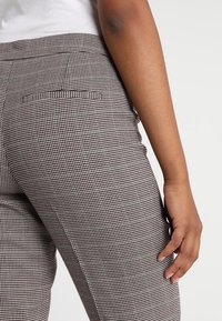 Bruna och vita houndstooth-mönstrade byxor med en figurnära design, som har en framficka och en slät yta. Midjebandet är synligt.