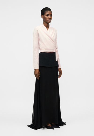 Kobieta ubrana w jasnoróżowy sweter na zakładkę zawiązany z boku oraz długą czarną spódnicę z falbaną i czarne buty na wskroś.