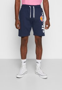Marineblaue Shorts mit einem Kordelzug, die auf der Seite mit einem weißen Logo bedruckt sind. Kombiniert mit weißen Turnschuhen und markierten Socken.