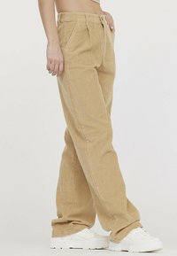 Pantalon en velours côtelé marron clair avec une coupe droite, doté de poches avant et d'un tissu texturé. Associé à des baskets blanches.