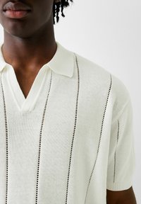 Polo s krátkým rukávem v světle krémové barvě s výstřihem do V. Disponuje vertikálními pruhy a malými výřezy, které přidávají texturu a detail.
