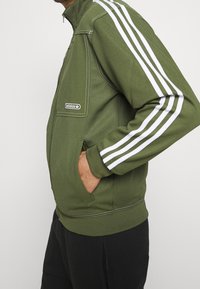 Grön zip-jacka med vit tre-stripes-design på ärmarna. Har en framficka och ribbade ärmslut. Tillverkad av texturerat tyg.