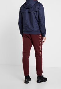 Marineblauwe hoodie met een rits aan de hals, gecombineerd met burgundy sweatpants met 'FC BARCELONA' gedrukt in lichtroze langs de zijkant. Zwarte sneakers.