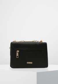 Czarna syntetyczna torebka na ramię z klapką, złotym łańcuszkiem, przednią kieszenią na suwak oraz małą metalową plakietką z logo. Teksturowane wykończenie i smukły kształt.