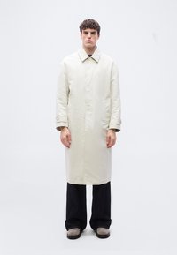 Νεαρός άνδρας όρθιος, φοράει μακρύ μπεζ παλτό, μαύρο φαρδύ παντελόνι και ανοιχτό γκρι παπούτσια μπροστά σε λευκό απλό φόντο.