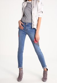 Sittande blå jeans med blommotiv broderade på vänster lår, i kombination med en randig skjorta och en ljus bomberjacka. Grå ankelboots.