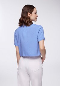 Camicia a maniche corte a righe blu e bianche con un design corto, dotata di colletto e tessuto morbido. Il modello indossa dei pantaloni rosa chiaro.