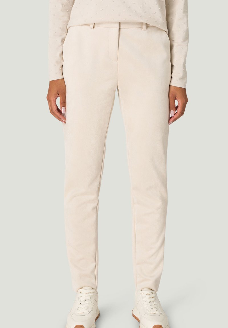 Beige tailored trousers made of soft fabric, featuring a flat front, side pockets, and a slim fit design. Worn with white sneakers.