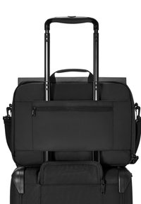 Czarna torba na laptopa zabezpieczona na wysuniętej rączce czarnej walizki na kółkach, z widocznymi kieszeniami na suwak i uchwytami do noszenia.