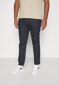 Jeans din denim albastru închis cu croială slim și design clasic cu 5 buzunare, asortați cu adidași albi cu logo-ul Nike auriu.