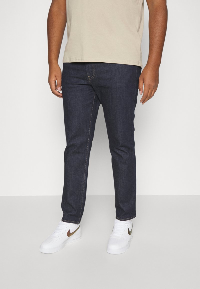 Jean en denim bleu foncé avec une coupe slim et un design classique à 5 poches, associés à des baskets blanches avec un logo Nike doré.