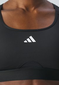 Zwarte sportbeha van soepel, rekbaar materiaal. Het heeft een ronde halslijn en een wit Adidas-logo aan de linkerkant.
