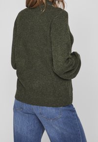 Olivgrön stickad tröja med hög krage, uppvikt ärm och en avslappnad passform, bärs över blå denimjeans.