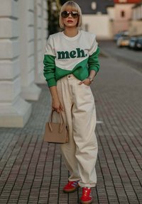 Zelená a bílá mikina s textem "meh.", krémové široké kalhoty, červené tenisky s modrými pruhy, sluneční brýle a krátké blond vlasy.