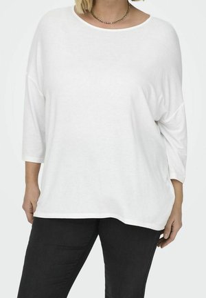 Valkoinen ylisuurikokoinen pitkähihainen paita pehmeästä kankaasta, jossa venepääntie ja laskostetut olkapäät, yhdistetty tummiin farkkuihin.