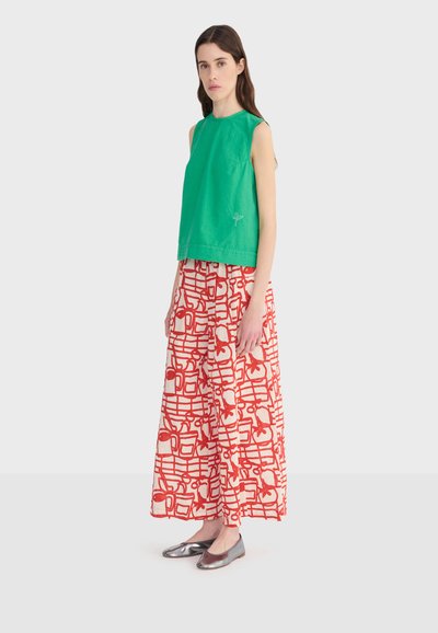 Žena odjevena u zeleni top bez rukava, široke crvene i bijele hlače s uzorkom te metalik srebrne ravne cipele, stoji na jednostavnoj pozadini.