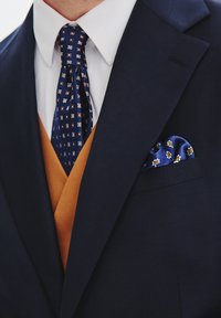 Granatowa marynarka z białą koszulą, złotą kamizelką i wzorzystym niebieskim krawatem. Chusteczka w kieszeni w kolorze niebieskim z czerwonymi i białymi akcentami. Gładka tekstura.