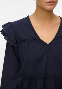 Blusa blu navy con accenti arricciati lungo le spalle, scollatura a V e balze arricciate in vita realizzata in un tessuto leggero.