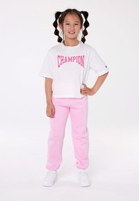 Camisa blanca de manga corta con texto rosa "CHAMPION" y pantalones de chándal rosa. El conjunto se completa con zapatillas deportivas blancas.