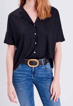 Femme portant une chemise noire à manches courtes boutonnée, rentrée dans un jean bleu avec une ceinture noire tissée avec une grande boucle ovale.