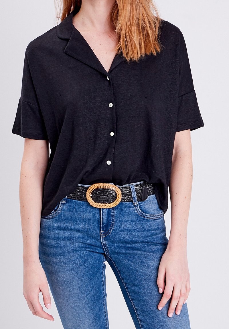 Femme portant une chemise noire à manches courtes boutonnée, rentrée dans un jean bleu avec une ceinture noire tissée avec une grande boucle ovale.