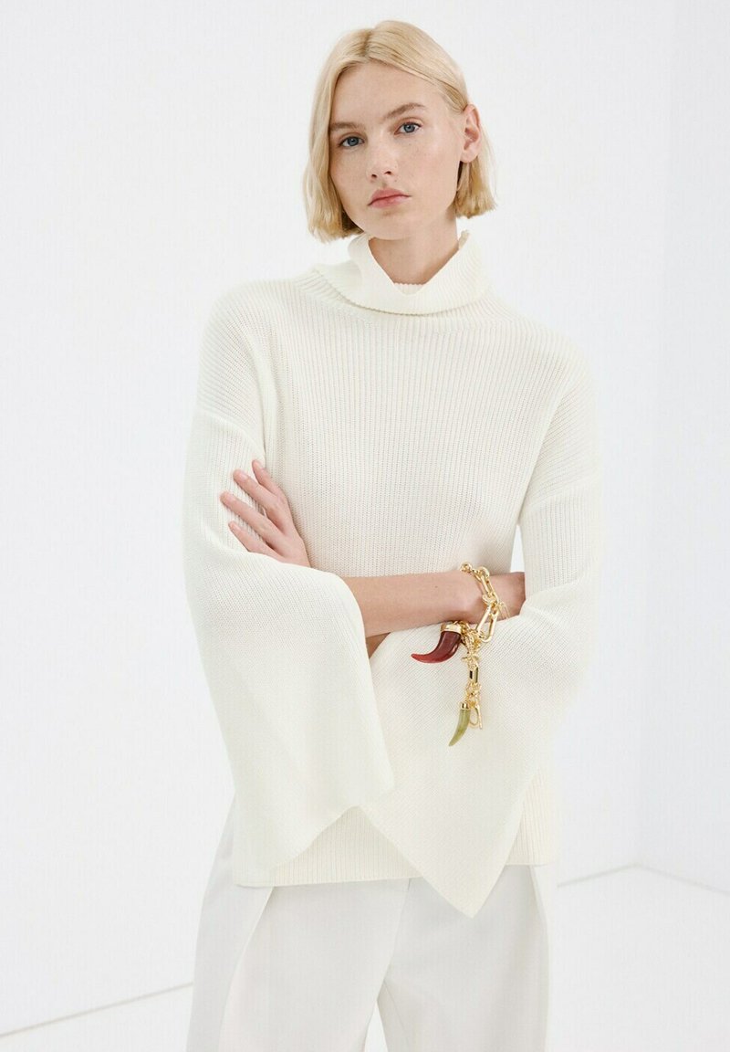 Plavolaska ženska s kratkimi lasmi nosi belo rebrasto rolko z razširjenimi rokavi, bele hlače in zlato ogrlico z obeski v rdeči in zeleni barvi.