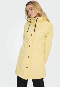 Rumena vodoodporna jakna z rdečo kapuco, črnimi vrvicami, pritisnimi gumbi in dvema sprednjima žepoma. Gladek, lahek material z udobnim krojem.