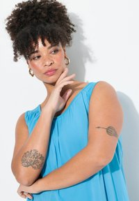 Ljusblå ärmlös klänning med rundad halsringning och veck. Modellen har en lockig uppsättning, guldörhängen i form av ringar och en blommig tatuering på sin arm.