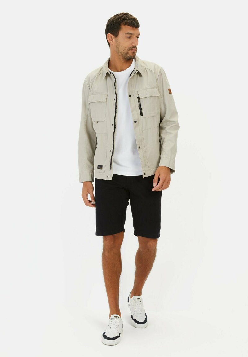 Lys grå jakke med lynlås og trykknap foran, fire lommer, parret med en hvid t-shirt og sorte shorts. Hvide sneakers med sorte detaljer.