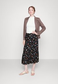 Blazer castanho sobre uma blusa branca, combinado com uma saia floral preta e sandálias de salto baixo bege. A saia apresenta um design com babados em camadas.