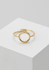 Une bague en or avec une forme ovale ouverte et une bande fine. Présente une finition lisse et un design minimaliste sur un piédestal blanc.