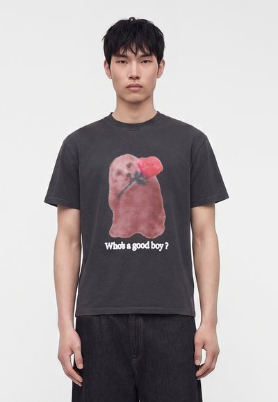 Jonge man draagt een zwart T-shirt met een hond die een rode bloem vasthoudt en de tekst "Who's a good boy?" op een effen witte achtergrond.
