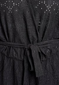 Vestido de encaje negro con un patrón texturizado y diseños de calados. Incluye un cinturón ajustable en la cintura para un ajuste ceñido. Detalle de caída sin costuras.